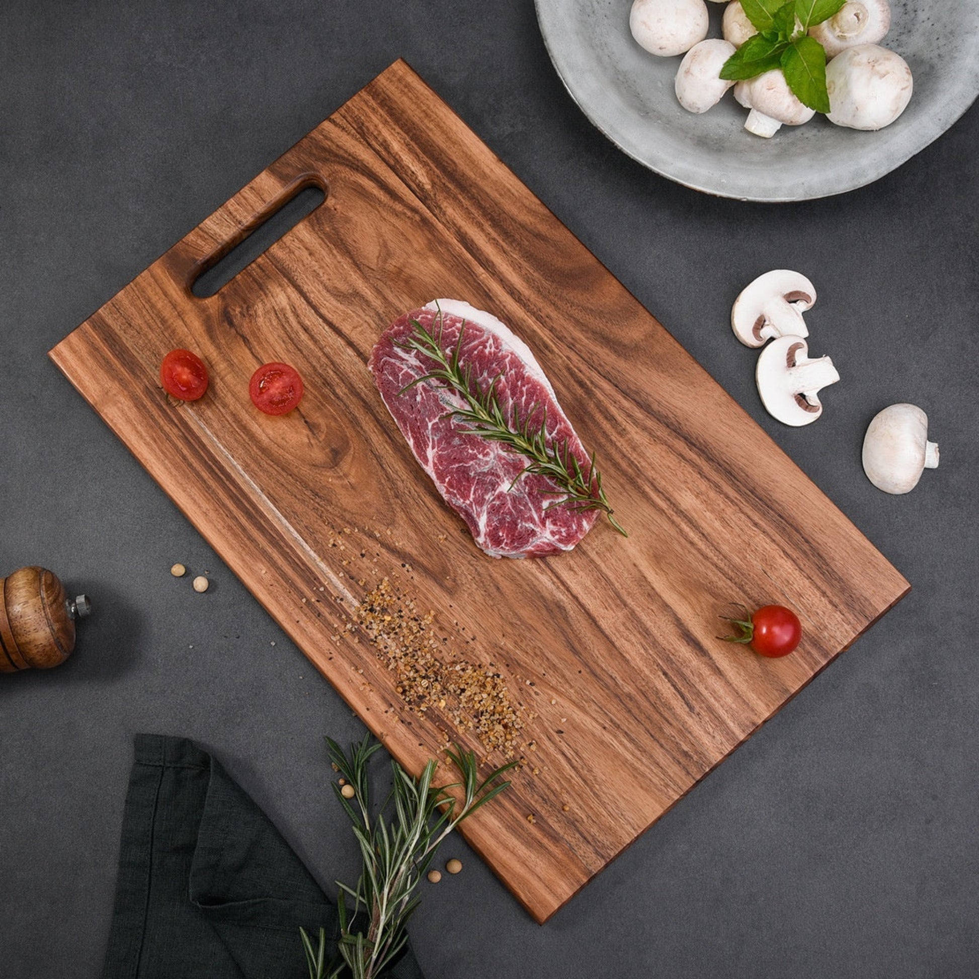 Raw steak on a wooden cutting board with vegetables and spices on a dark grey kitchen surface. Wooden acacia mangium cutting board with a handle. Dark grey surface. Earthy, warm and natural ambience. 