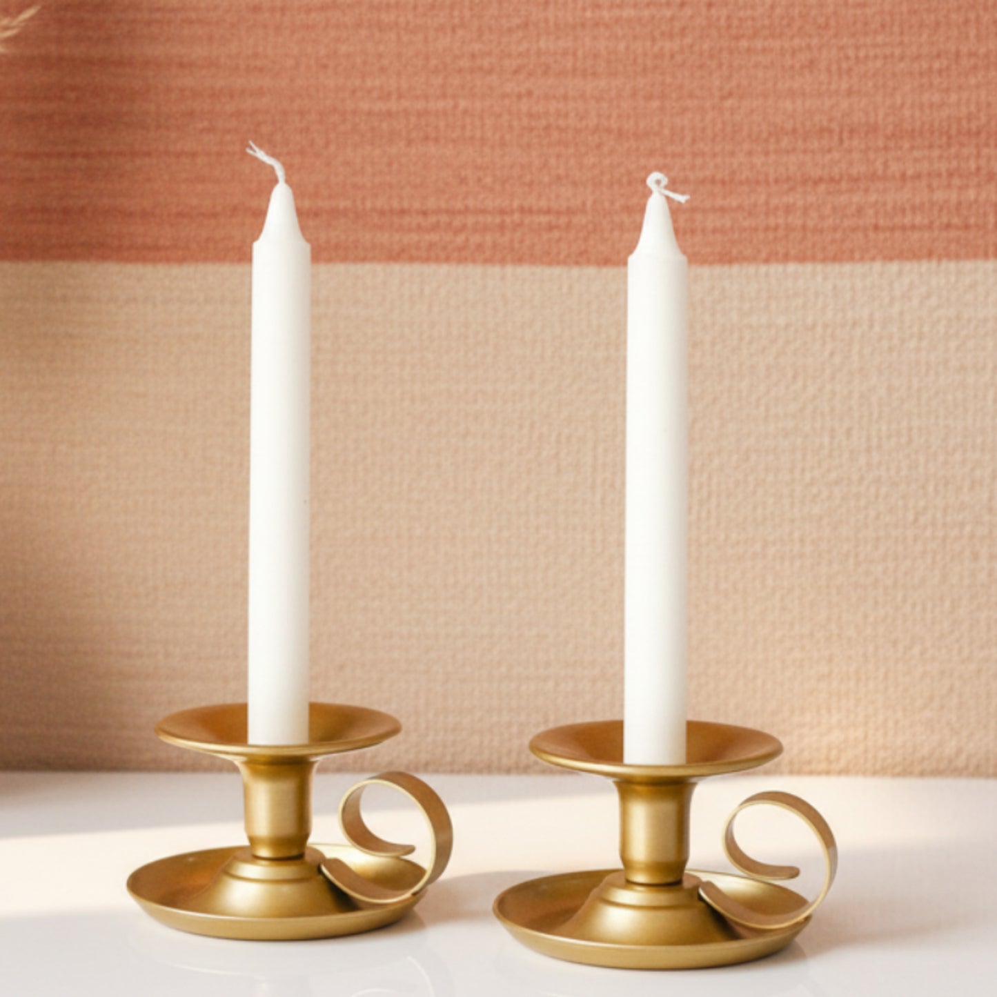 Two gold candle holders with white candles on a white surface with a warm and earthy background. Minimalist design. Elegant, modern and antique. 