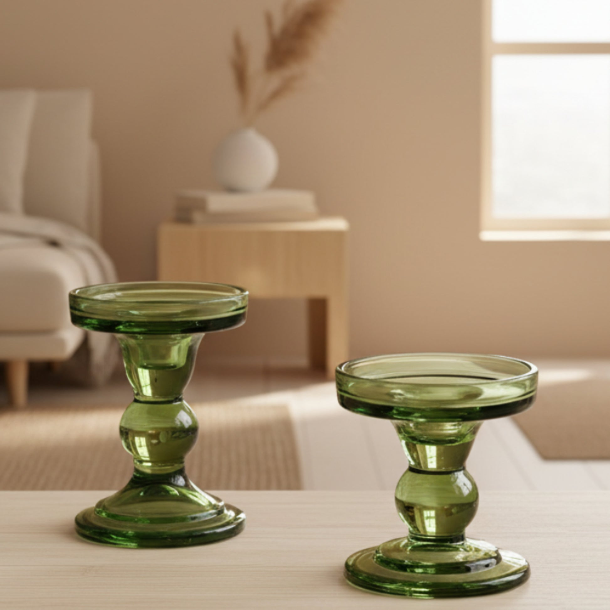 Two green glass candle holders on a wooden table with a beige-brown blurred earthy background. Sofa, dried flowers, warm and earthy atmosphere.