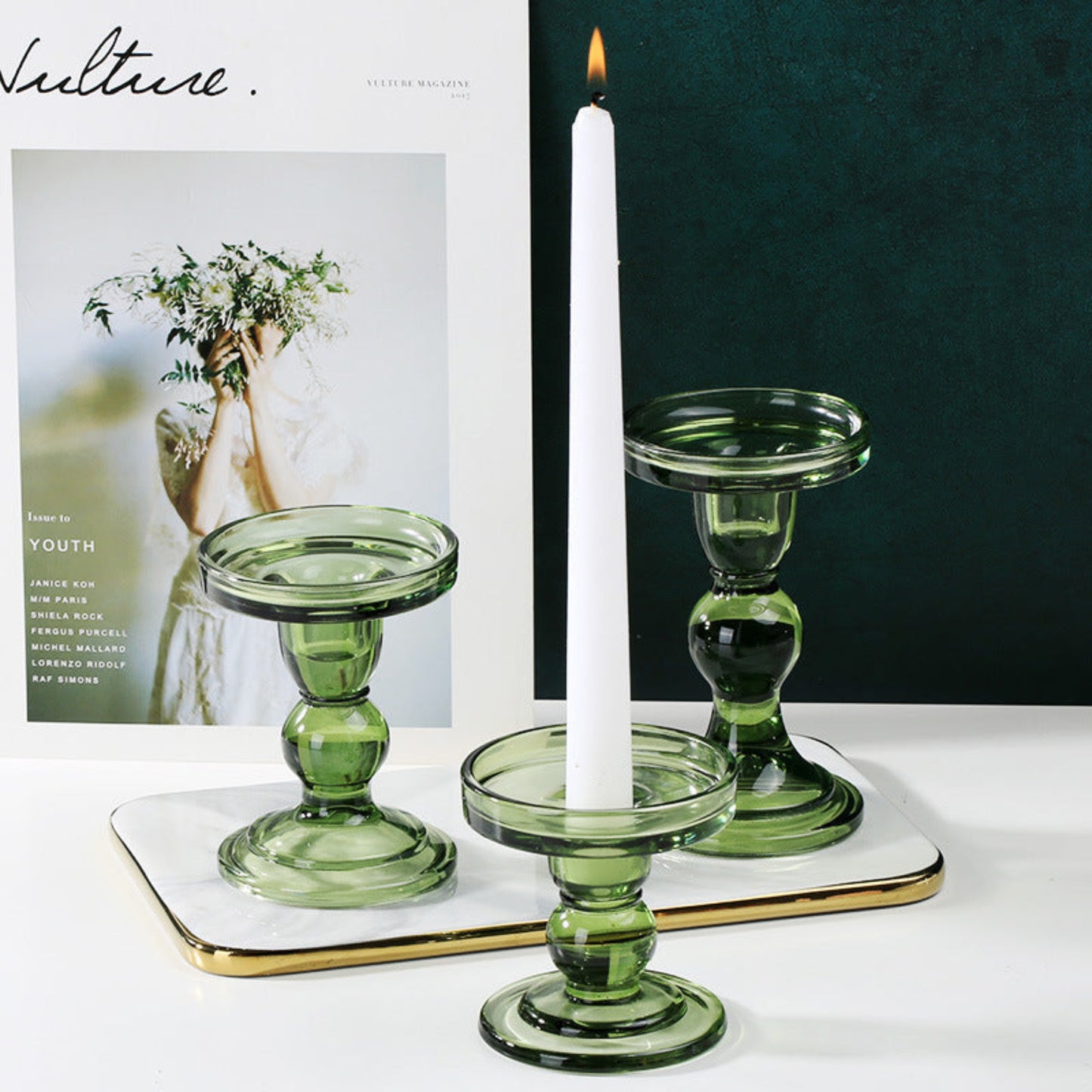 Three green glass candle holders, one with a white candle, in front of a magazine cover background on a white surface. Green wall background on the right. 