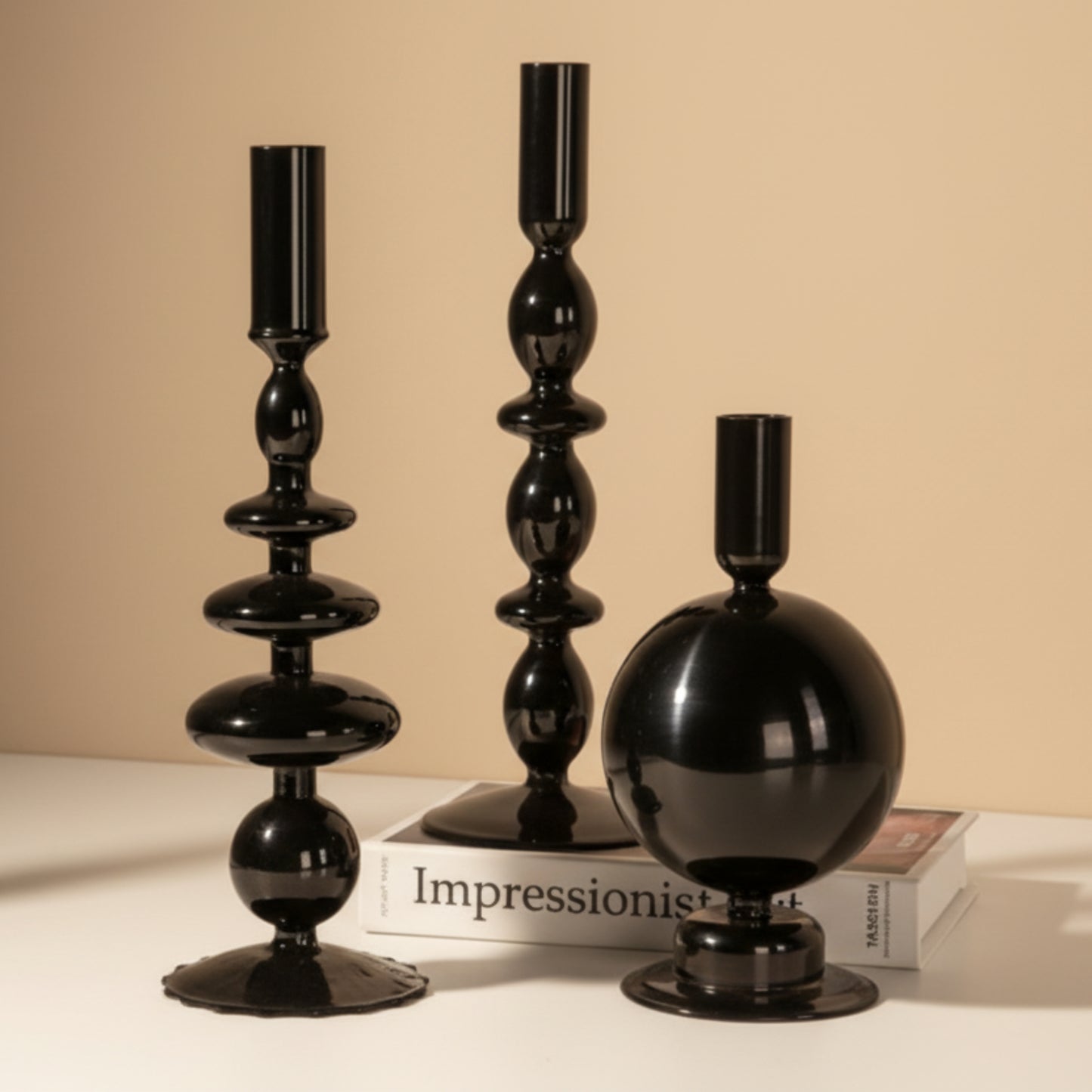 Set of three black decorative candle holders on a white surface with a book and on a beige warm background. 
Scandinavian inspired minimalist decor design. Sleek and modern decor. 