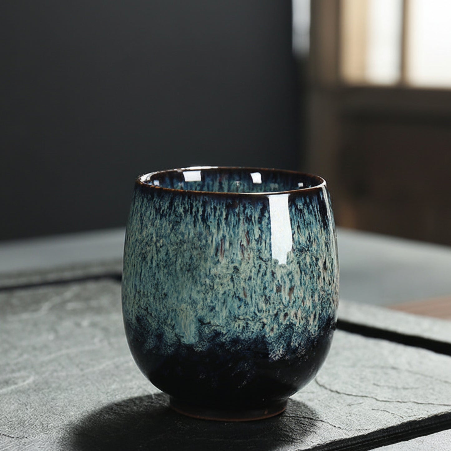 Blue ceramic cup with unique glaze pattern on a textured surface and a blurred background. Natural and earthy ambience. 