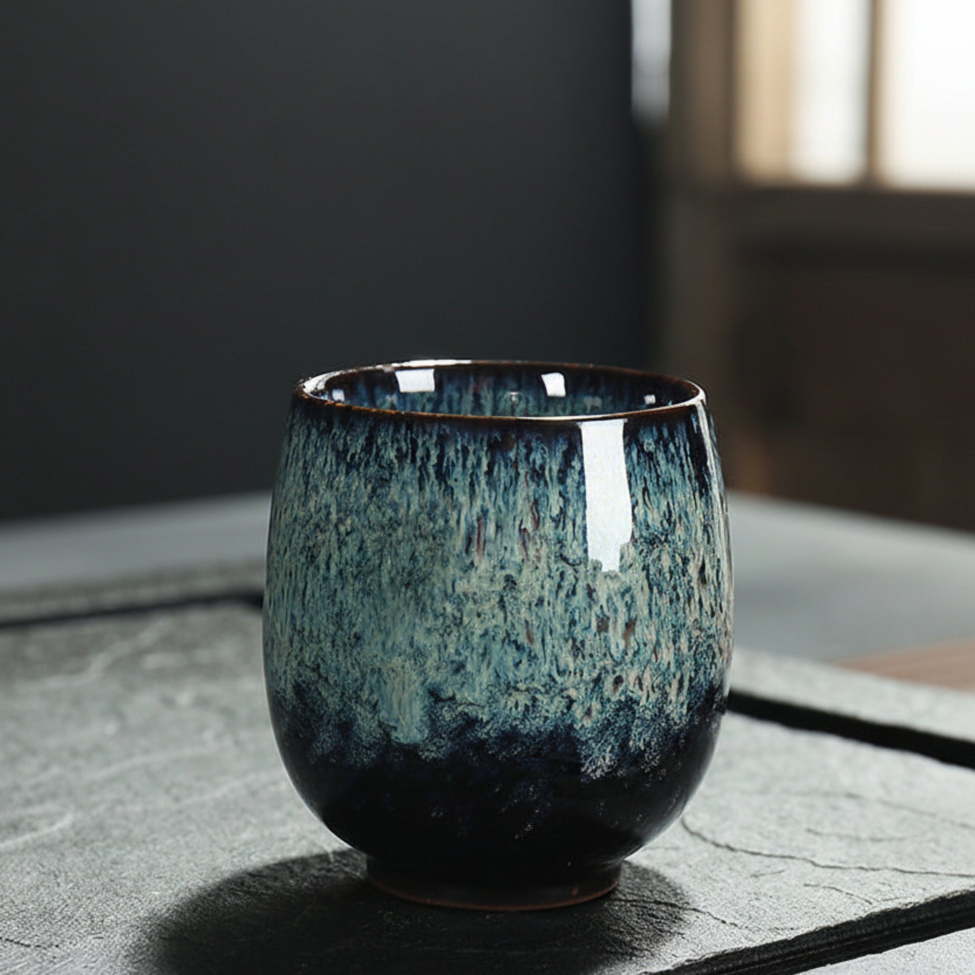 Blue ceramic cup with unique glaze pattern on a textured surface and a blurred background. Natural and earthy ambience. 