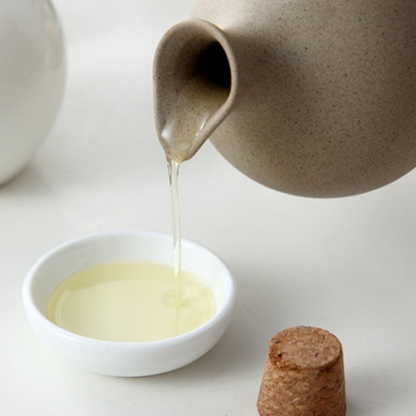 Ceramic jar pouring liquid into a small white bowl on a light background. 
Beige ceramic oil dispenser with cork lid on a light surface. 