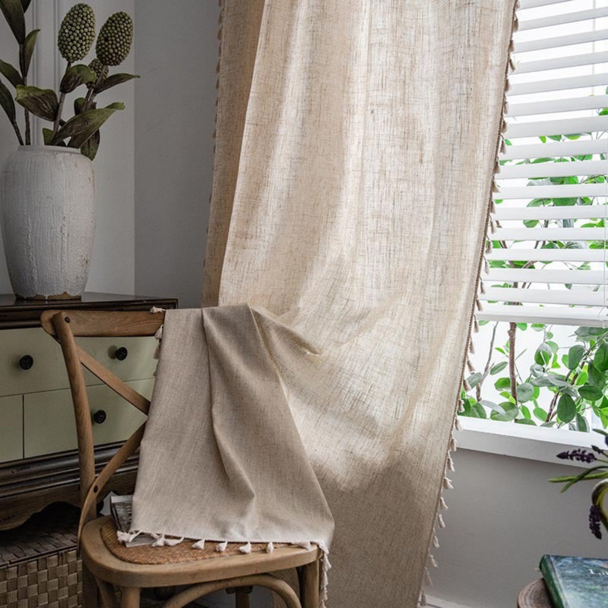 Beige curtain draped over a chair with a plant and window in the background. Natural decor styling curtain for calm and modern interiors.