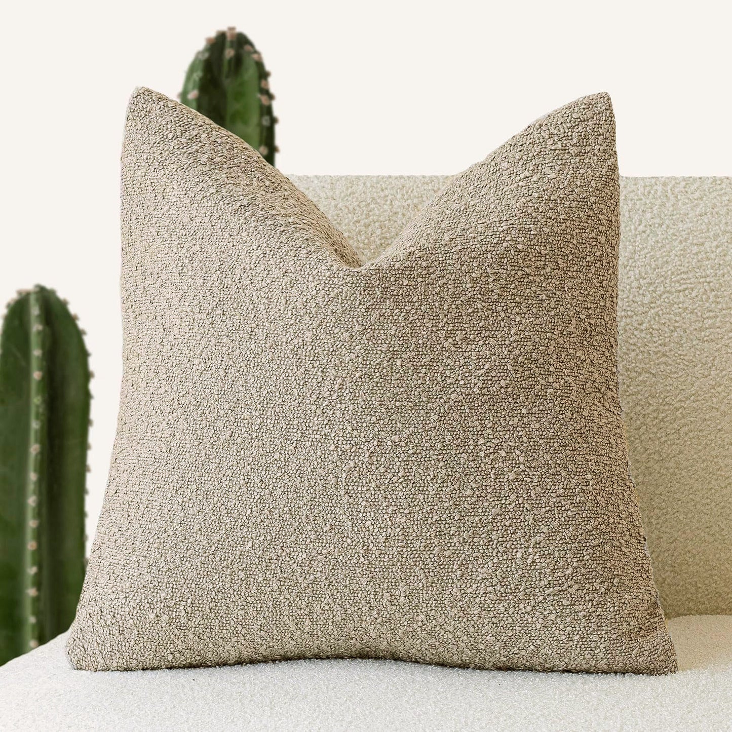 A beige Decorative Boucle Pillow Cover with a textured surface displayed on a neutral background and a cactus in the background. Warm and cozy decor ambience. 
