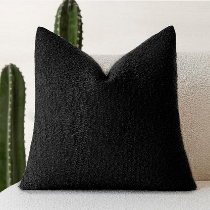A black Decorative Boucle Pillow Cover with a textured surface displayed on a neutral background and a cactus in the background. Warm and cozy decor ambience. 