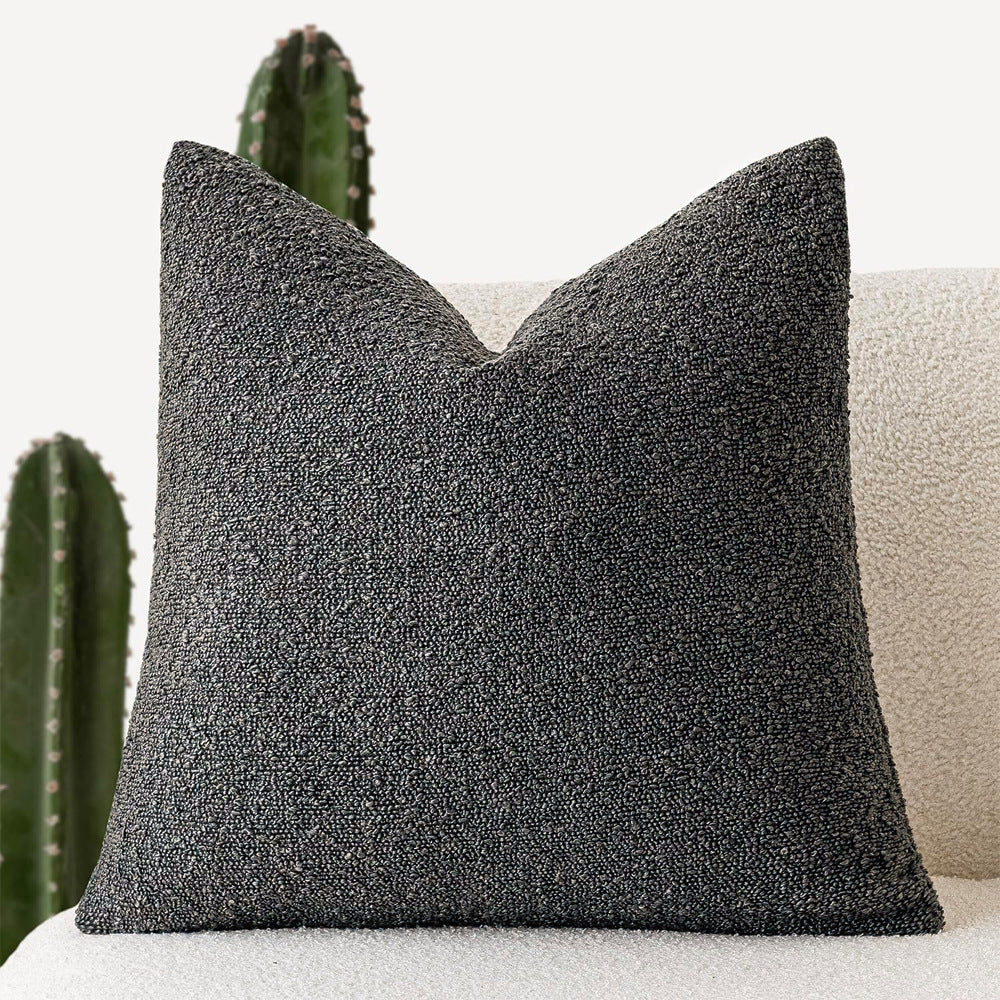 A dark gray Decorative Boucle Pillow Cover with a textured surface displayed on a neutral background and a cactus in the background. Warm and cozy decor ambience. 