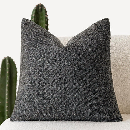 A dark gray Decorative Boucle Pillow Cover with a textured surface displayed on a neutral background and a cactus in the background. Warm and cozy decor ambience. 
