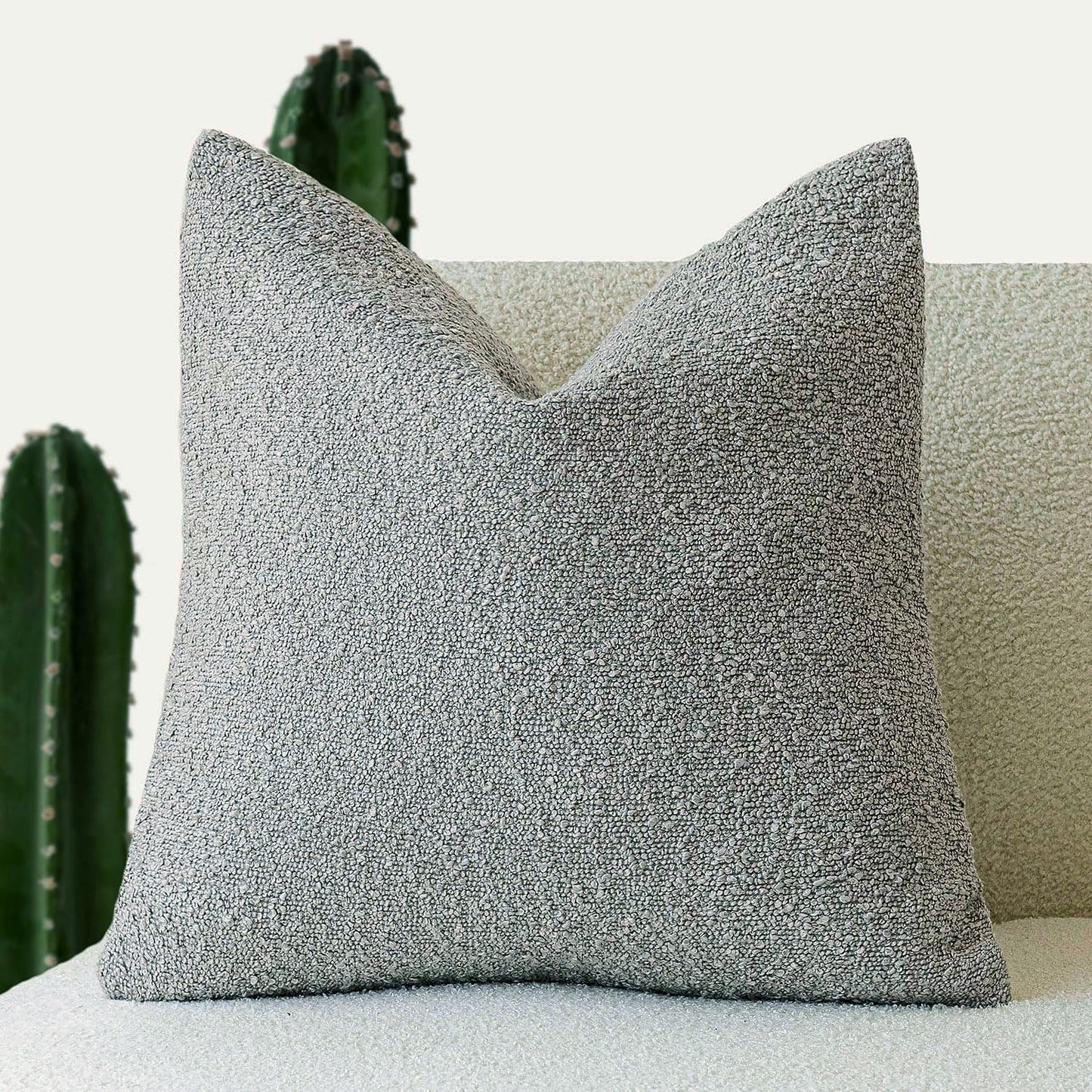A gray Decorative Boucle Pillow Cover with a textured surface displayed on a neutral background and a cactus in the background. Warm and cozy decor ambience. 