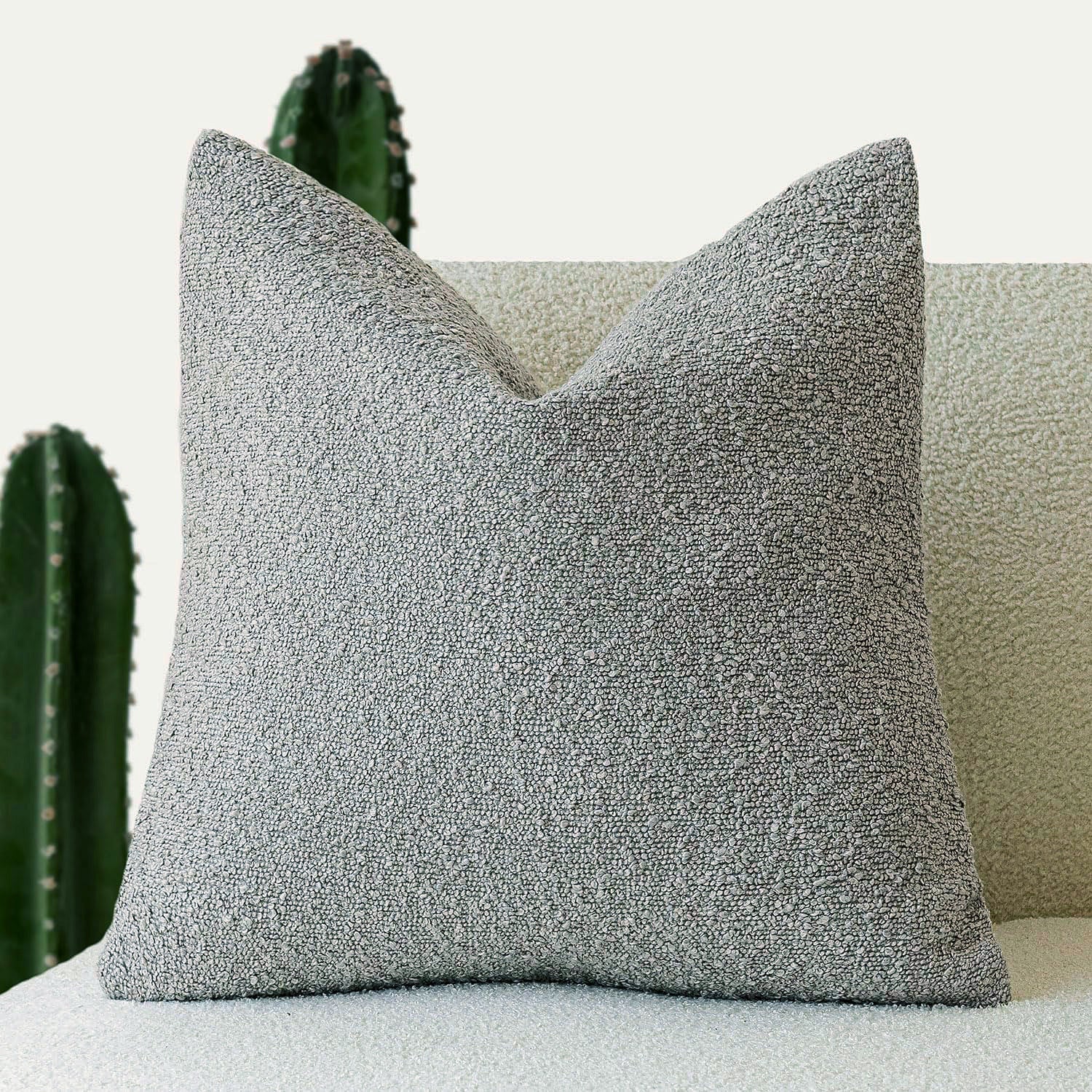 A gray Decorative Boucle Pillow Cover with a textured surface displayed on a neutral background and a cactus in the background. Warm and cozy decor ambience. 