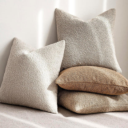 Four textured boucle pillows in beige, brown, and gray on a natural background. Warm and cozy decor ambience. 