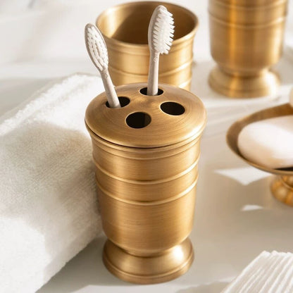Toothbrush holder with two toothbrushes in it on a white surface together with other items of the collection and a white towel. A set of gold-colored copper bathroom accessories. Stylish golden bathroom decor designed for every bathroom space. 