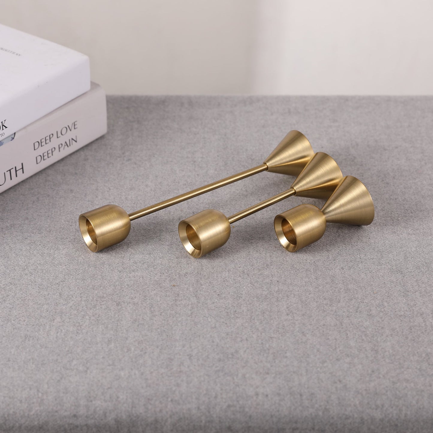 Set of three gold candle holders with different sizes on a grey surface and a white background. Two books in the picture. Minimalist home decor. Timeless design. Modern home decor.  