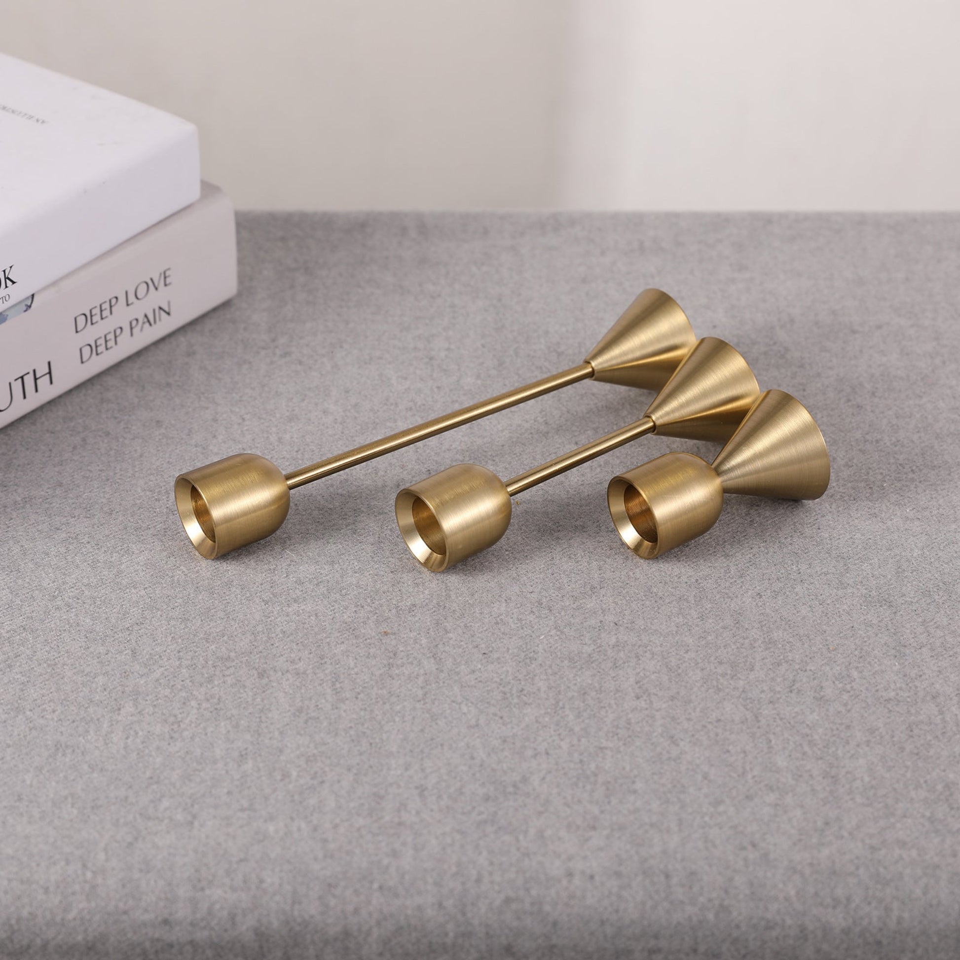 Set of three gold candle holders with different sizes on a grey surface and a white background. Two books in the picture. Minimalist home decor. Timeless design. Modern home decor.  