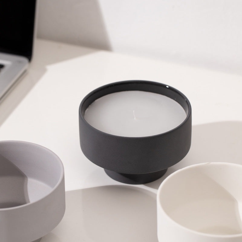 Set of three ceramic bowls in different colors on a white surface on a white wall background with a laptop in the picture. Scandinavian decor simplicity. Minimalist handmade scented candles. Black candle is in the center. 