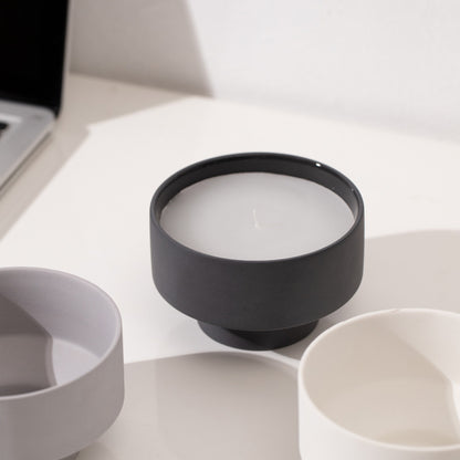 Set of three ceramic bowls in different colors on a white surface on a white wall background with a laptop in the picture. Scandinavian decor simplicity. Minimalist handmade scented candles. Black candle is in the center. 