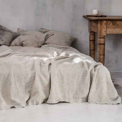 Bed with light gray plain linen bed sheet and pillows in a minimalistic earthy room on a grey background, with a wooden table in the background. Earthy and natural home and bed decoration. 