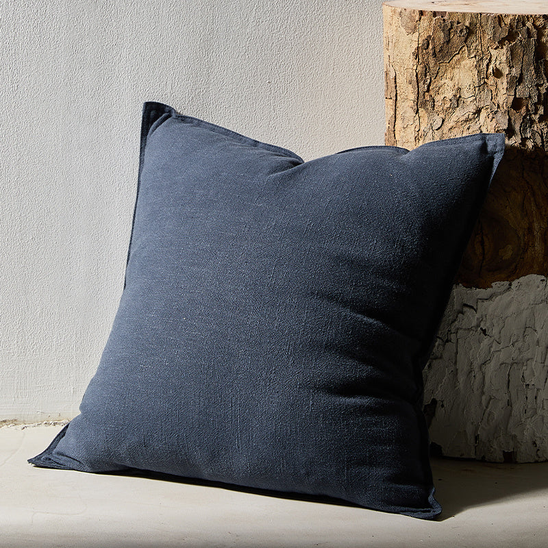 A blue pure cotton and linen pillow on a earthy surface with a plain white wall in the background. Warm and natural ambience. Wood in the background. 