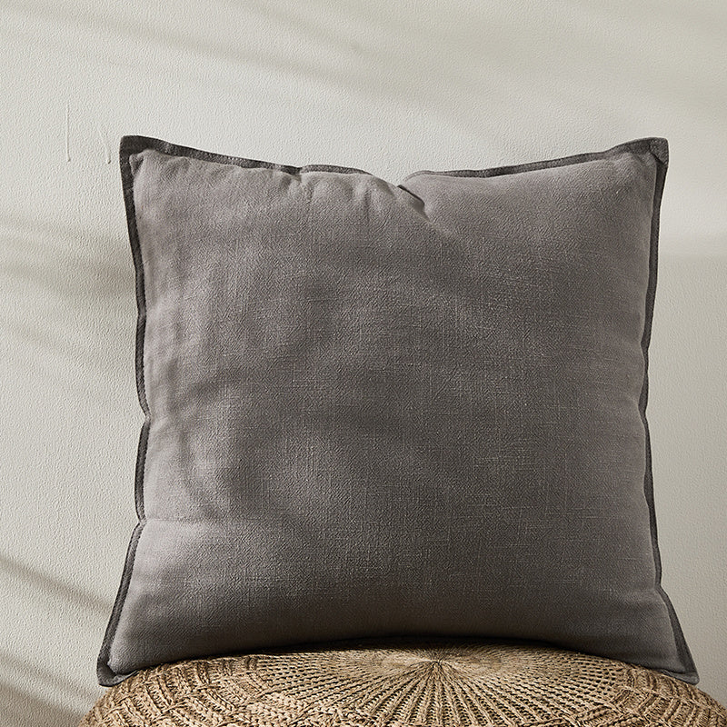 A grey pure cotton and linen pillow on a natural surface with a plain white wall in the background. Warm and natural ambience. 