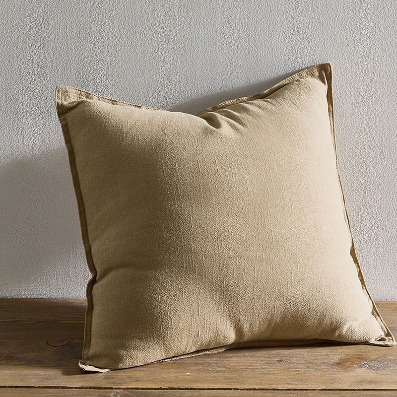 A beige pure cotton and linen pillow on a wooden surface with a plain white wall in the background. Warm and natural ambience. 