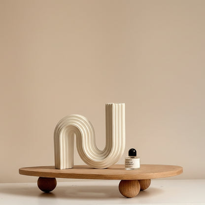 Wooden tray with unique ball high foot design on a white-beige surface and against a white beige wall. Natural and earthy decor item with a irregular triangle shape that holds a parfume and another decor item. 
