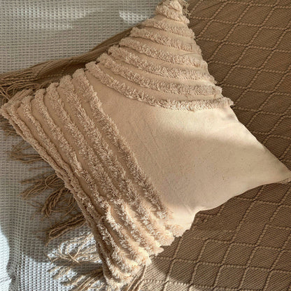 Beige textured pillow on a blanket with tassels and on a white fabric textured surface. Scandinavian interior with warm light.  