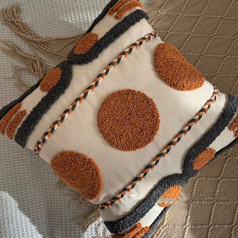 Beige, black and orange textured pillow on a blanket with tassels and on a white fabric textured surface. Scandinavian interior with warm light. Decorative pillow with brown circular patterns. 