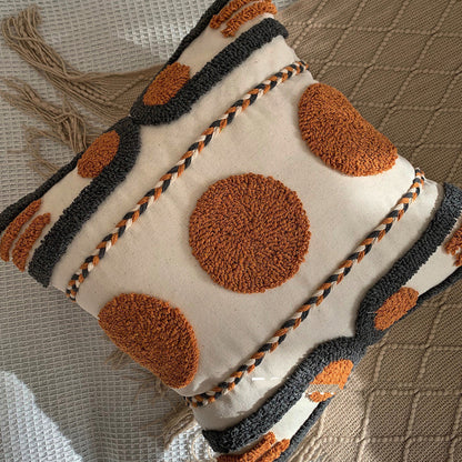 Beige, black and orange textured pillow on a blanket with tassels and on a white fabric textured surface. Scandinavian interior with warm light. Decorative pillow with brown circular patterns. 