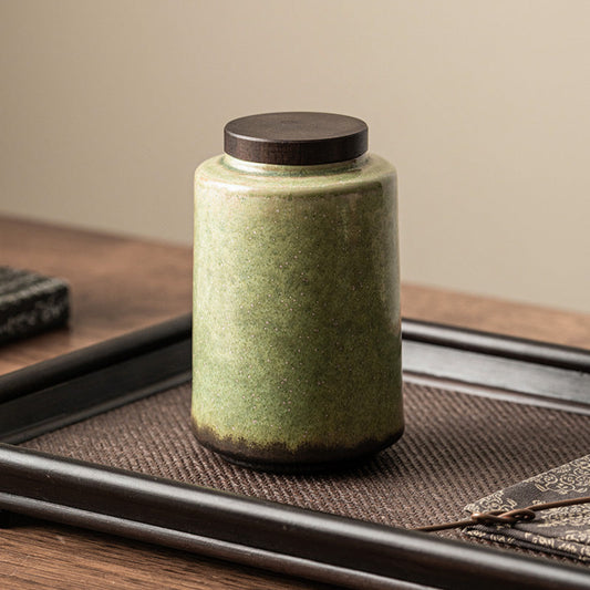 Green ceramic jar with a dark brown lid on tray and a wooden surface on a beige wall background. Earthy home decor with natural textures and warm tones. Timeless and minimalist ceramic for kitchen. 