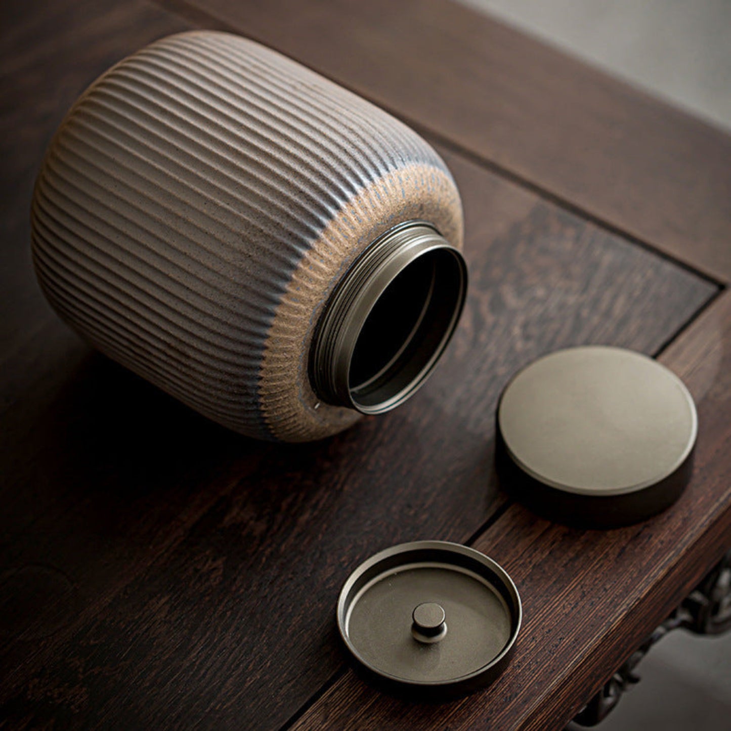 Textured ceramic container with open metal lid on a wooden table surface. Grey-brown earthy and warm kitchen item. Minimalist home decor and tea storage. 