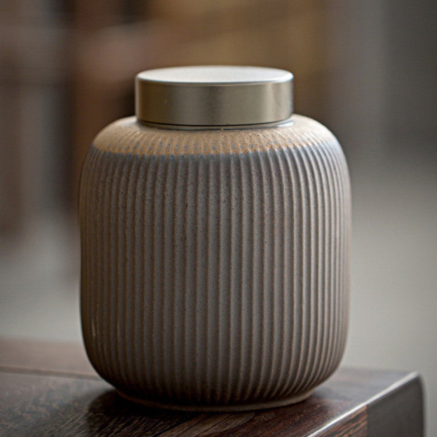 Ceramic jar with ribbed texture and metallic lid on a wooden surface against a blurred background. Grey-brown earthy and warm kitchen item. Minimalist home decor and tea storage. 