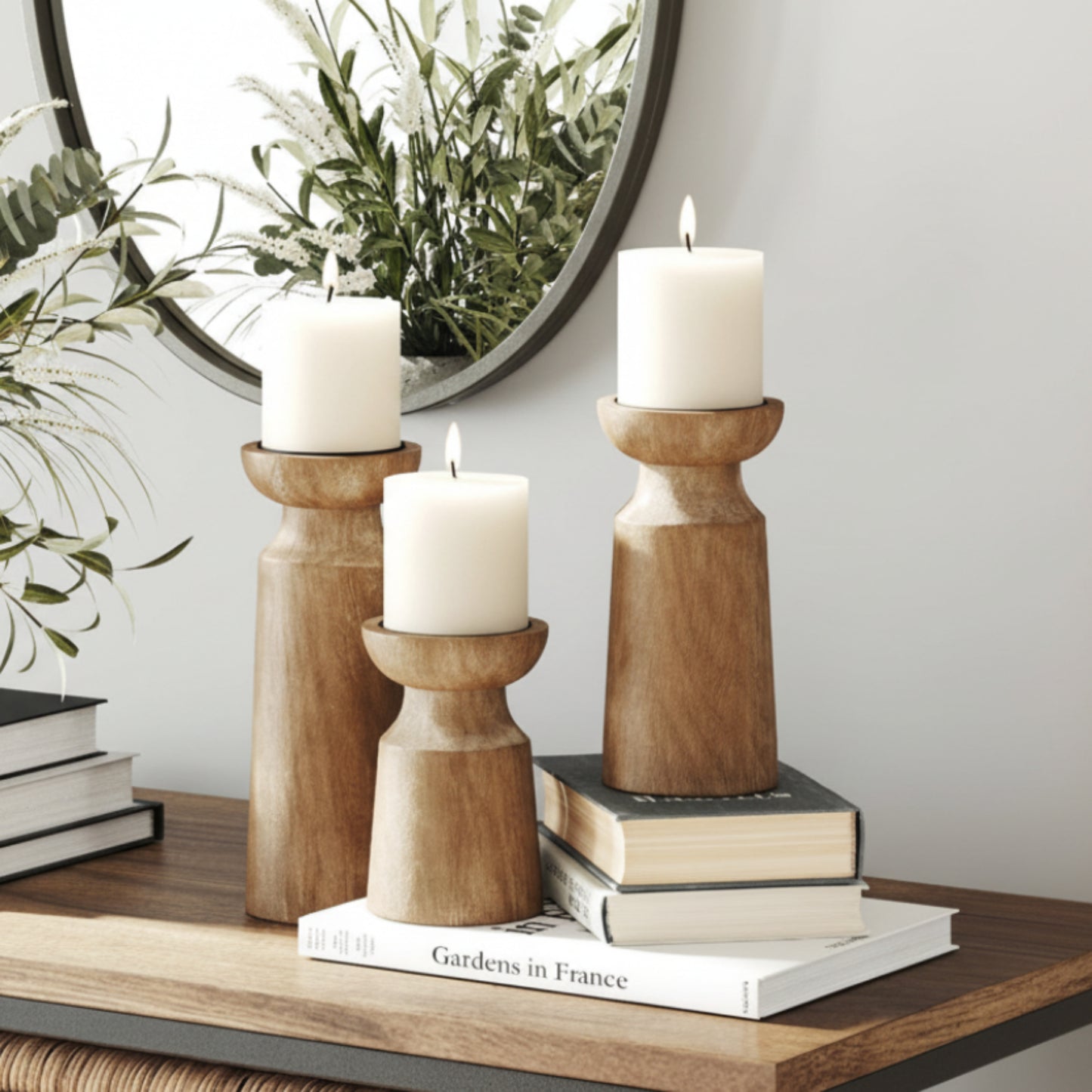Three wooden candle holders with candles on a wooden shelf and a white wall background. Two out of three wooden candle holders stand on books. A mirror in the background.